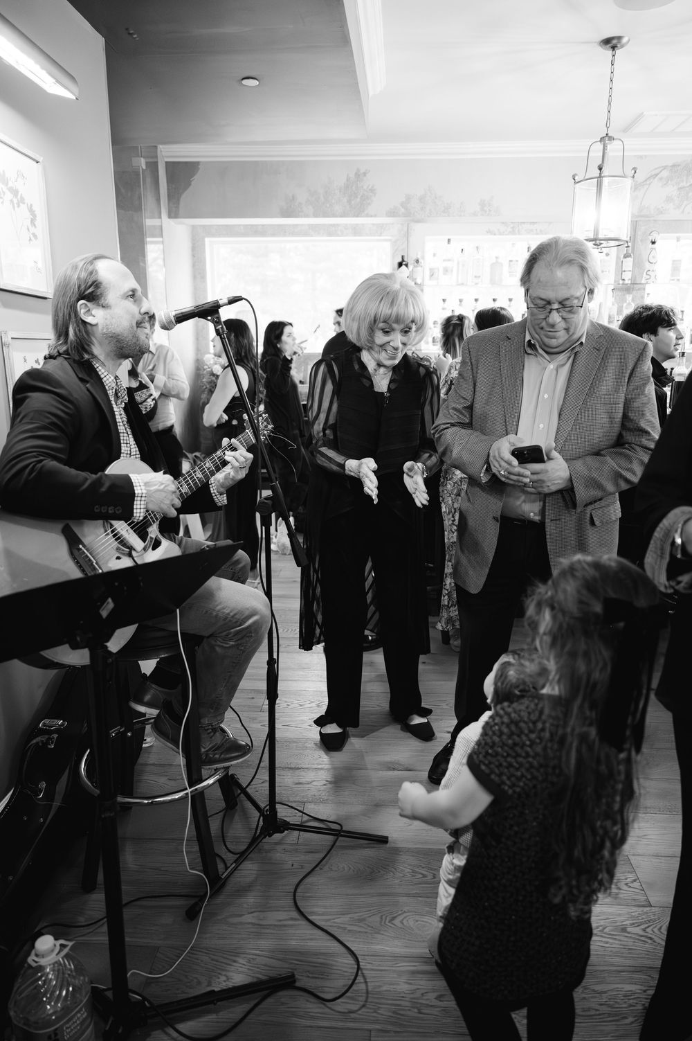 Live guitarist performs at an indoor event as guests mingle nearby in black and white photo.
