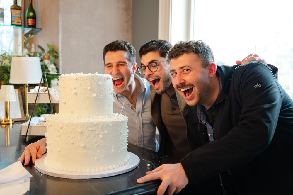 Three excited men pose with a white two-tier wedding cake at a celebration event.