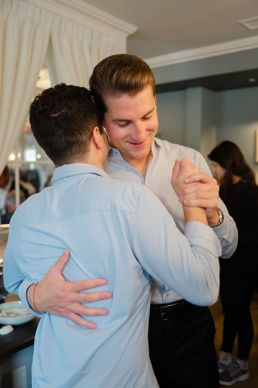 Two men in light blue dress shirts share a dance together, smiling warmly at an indoor event.