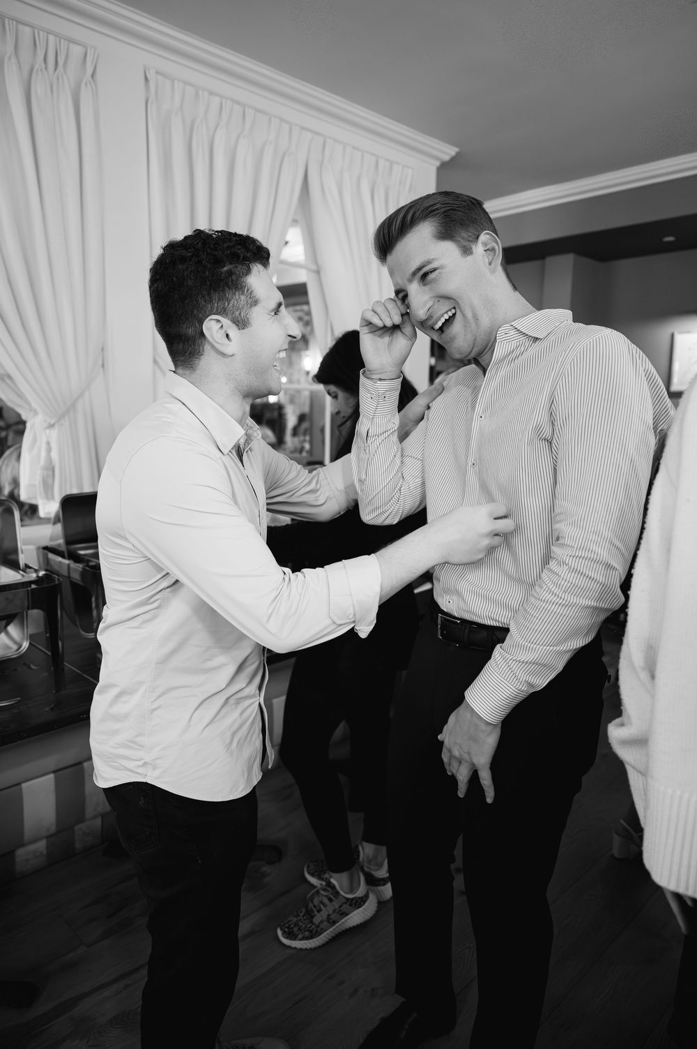 Two men laughing and adjusting shirts in black and white wedding preparation photo.