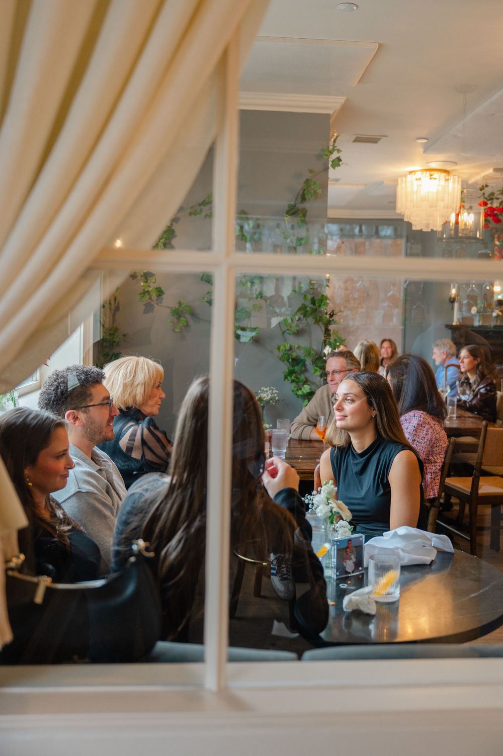 Guests dining inside an elegant restaurant with green ivy walls and chandelier, photographed through a window.