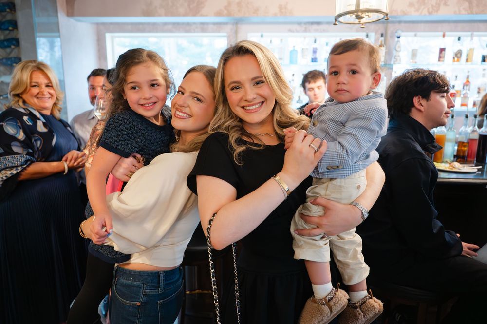 Two women smile holding children at a festive indoor family gathering.
