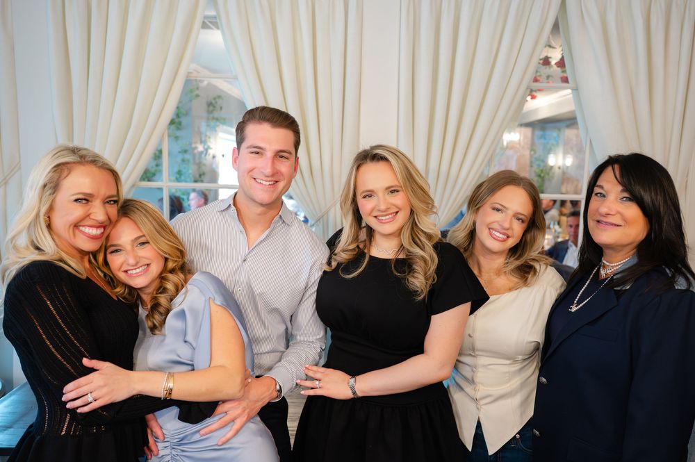Group of six smiling people posing together in a bright elegant restaurant with cream curtains.