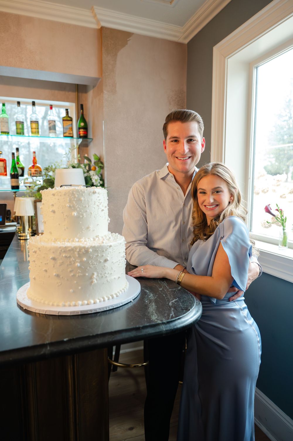 Smiling couple posing beside elegant two-tier white wedding cake at bar counter.