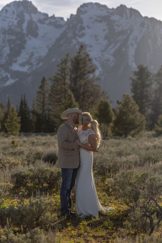 Couple embracing in a meadow with snow-capped mountains, groom in cowboy hat, bride in white gown, outdoor wedding.