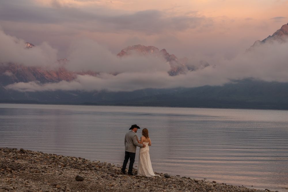 Couple in wedding attire embracing on rocky lakeshore at sunset with dramatic pink clouds and mountain backdrop.