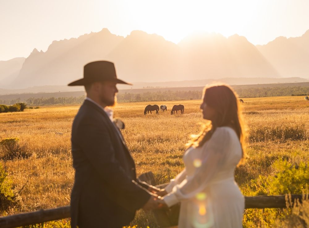 Couple holds hands at golden sunset with mountain backdrop and horses grazing in a scenic field.