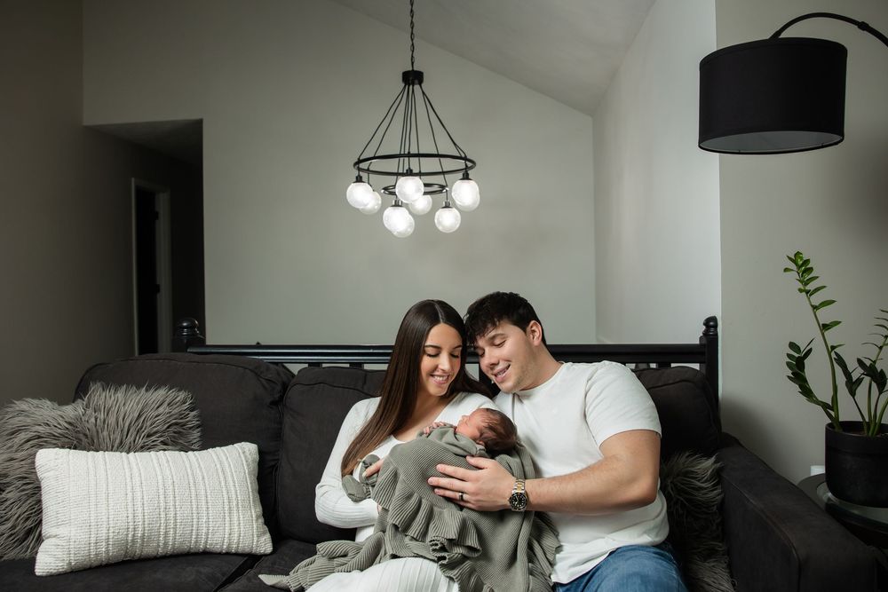Young couple sitting on gray sofa holding newborn baby wrapped in green blanket at home.
