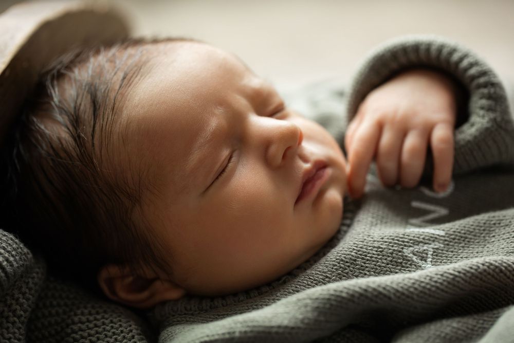 Serene close-up of sleeping newborn baby face with dark hair in sage green knit wrap.