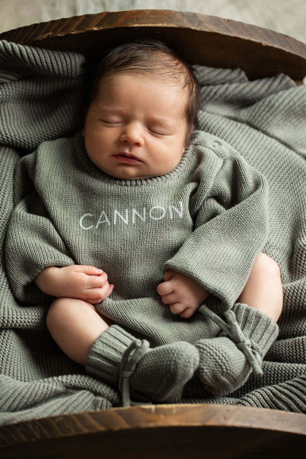 Newborn baby sleeping in wooden bowl wearing sage green knit Cannon personalized romper.