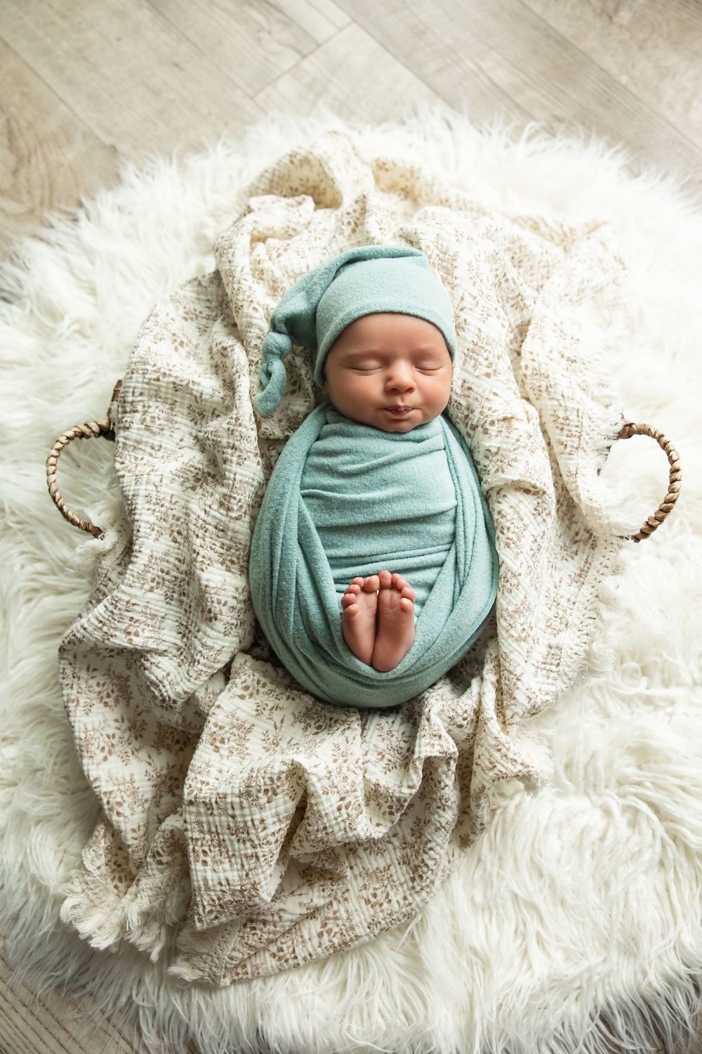 Swaddled newborn baby in teal wrap and knotted hat sleeping in basket on white fur.
