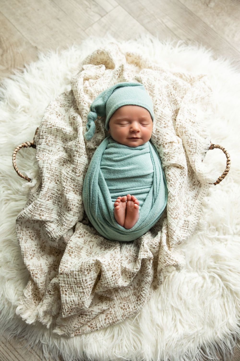 Top view of swaddled newborn in teal wrap and hat sleeping in wicker basket on fur rug.