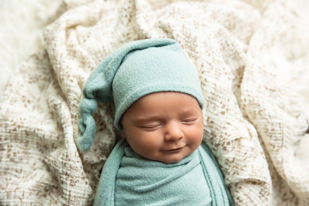 Smiling sleeping newborn baby swaddled in teal wrap wearing matching knotted sleep hat.