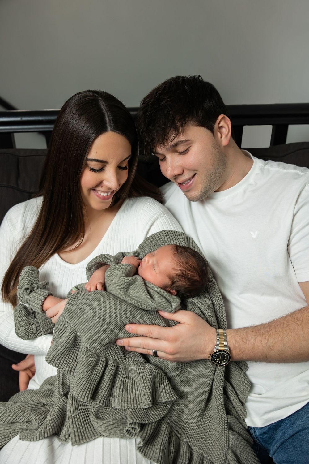 Smiling new parents holding sleeping newborn baby wrapped in green blanket during newborn session.