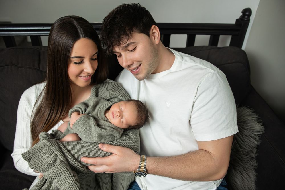 New parents cuddling newborn baby wrapped in sage green blanket during in-home photo session.