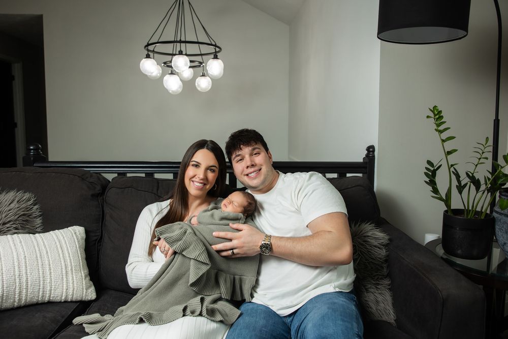 Happy new parents sitting on couch holding newborn baby wrapped in green blanket at home.