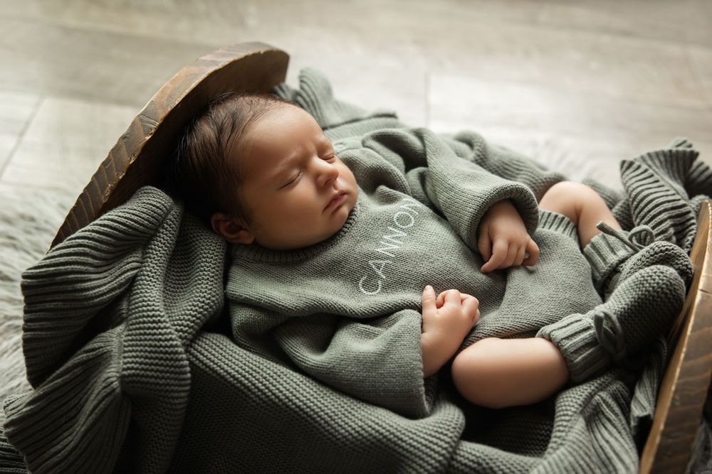 Sleeping newborn baby in sage green knit Cannon outfit nestled in wooden bowl with matching blanket.