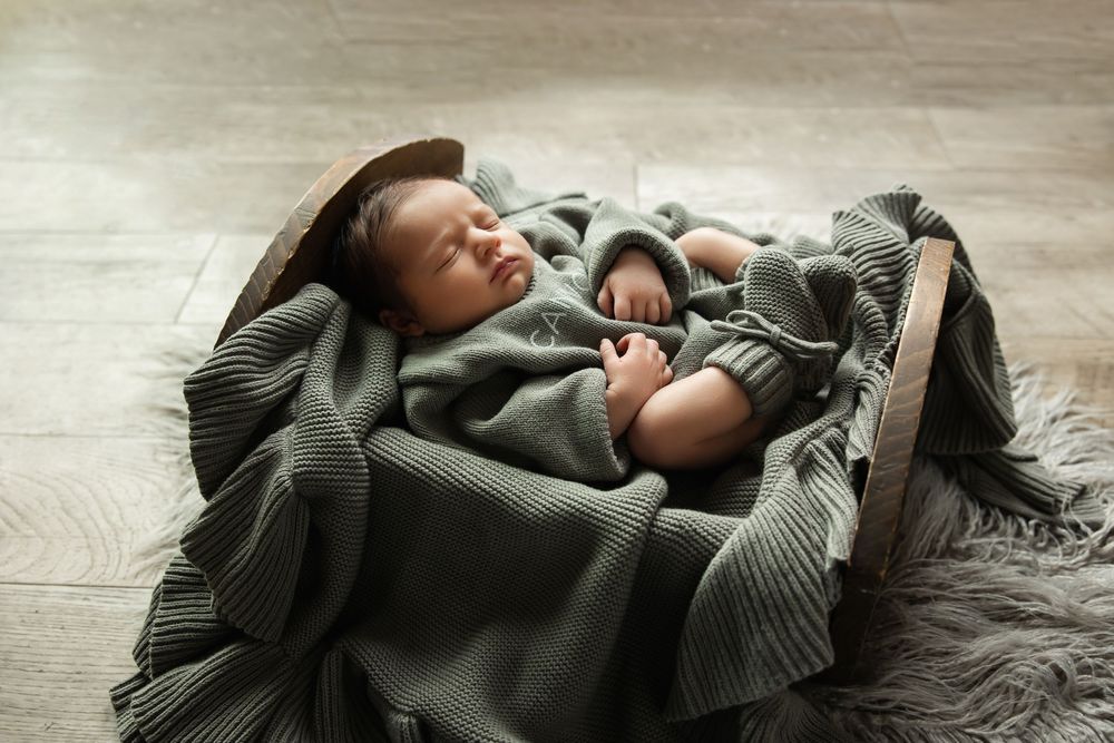 Peaceful newborn baby wrapped in sage green knit blanket sleeping in rustic wooden bowl prop.