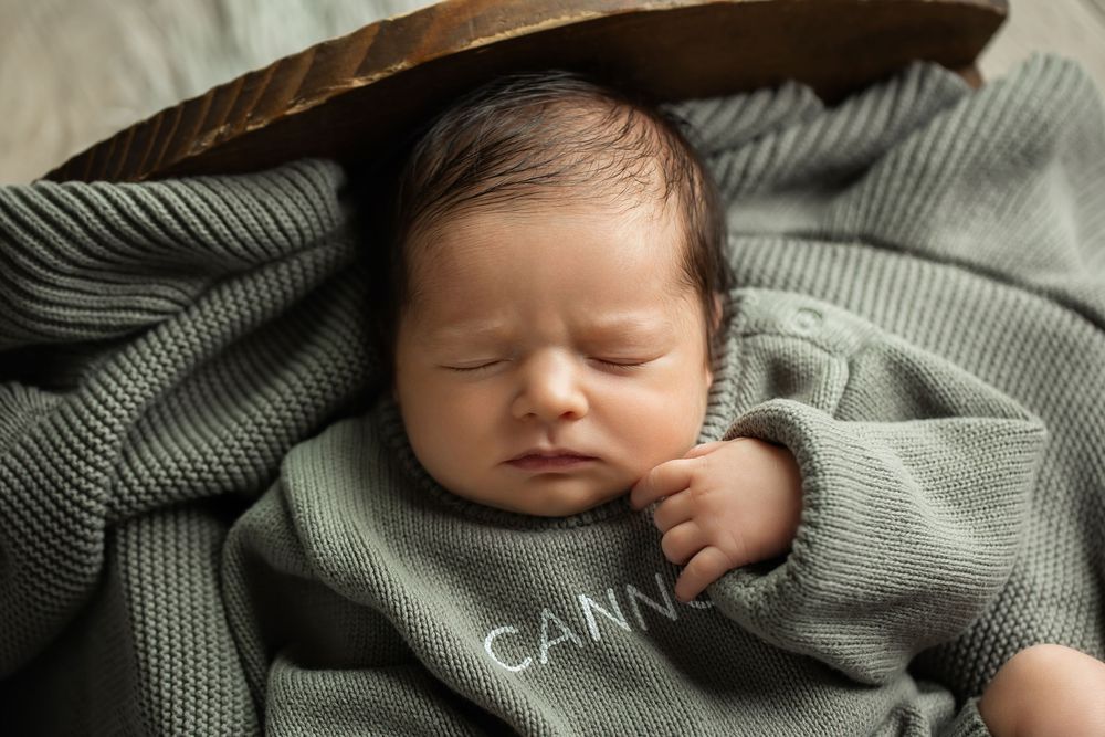 Close-up of sleeping newborn in sage green Cannon knit romper resting in wooden bowl.