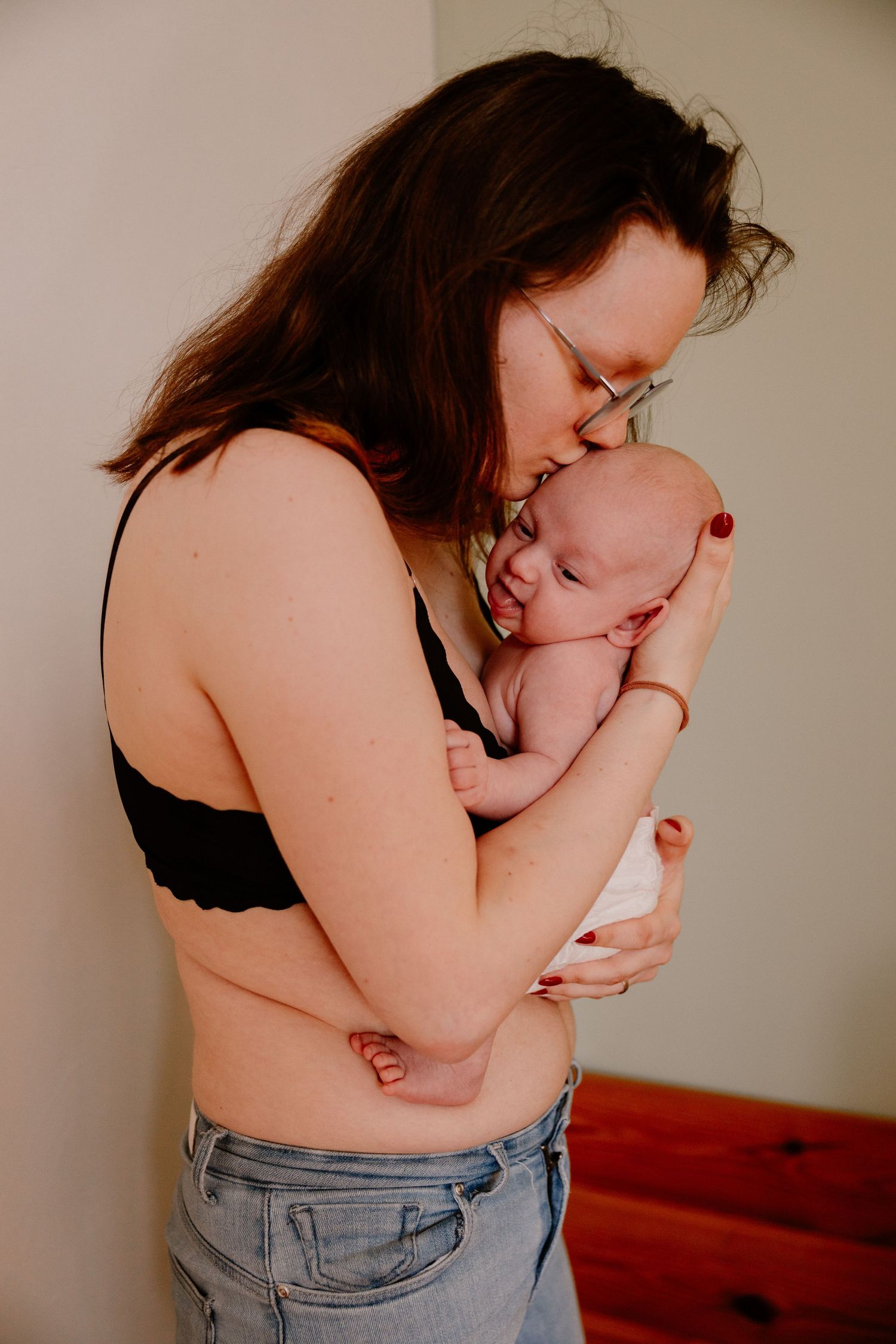 Mother in black bralette and jeans tenderly kissing her newborn baby while standing indoors.