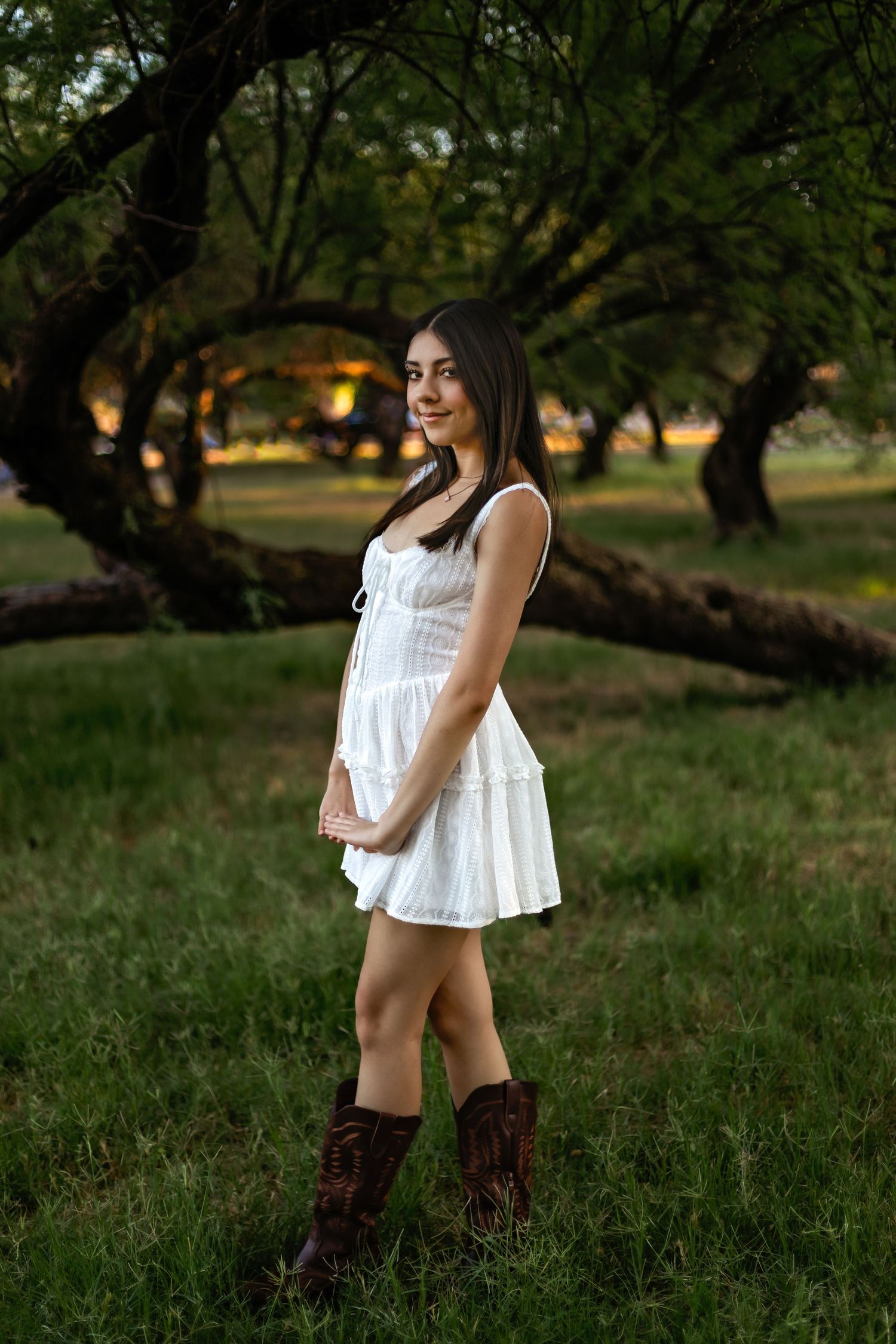 Young woman in white sundress and brown cowboy boots smiling in a green park at dusk.