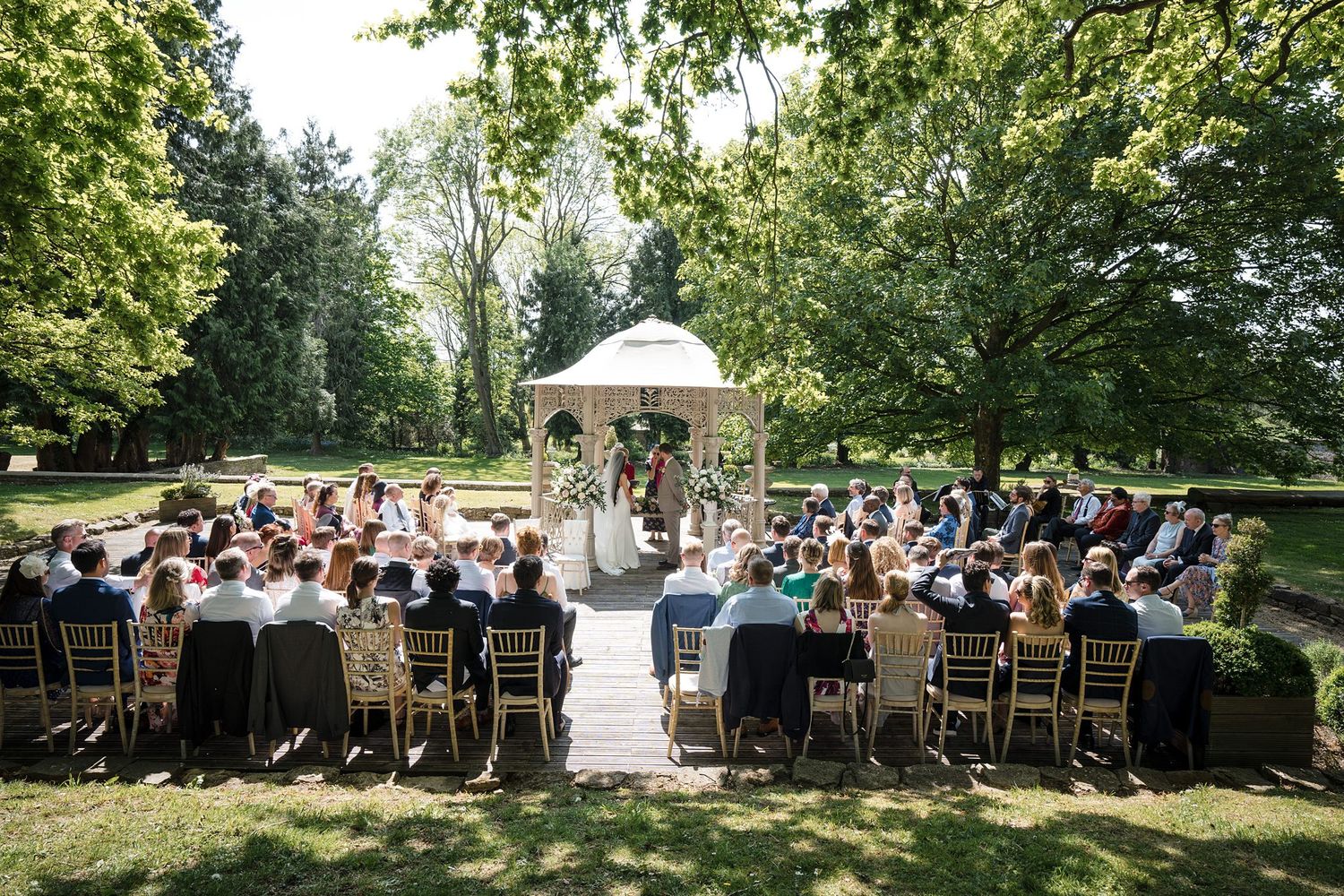Outdoor wedding ceremony with guests seated on gold chairs facing a gazebo surrounded by lush green trees.