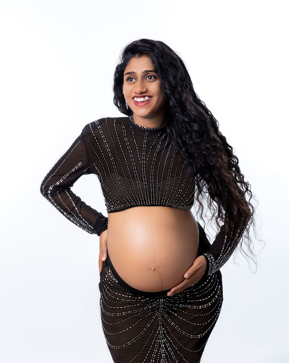 Pregnant woman in black beaded crop top and skirt cradling baby bump, smiling against white background.