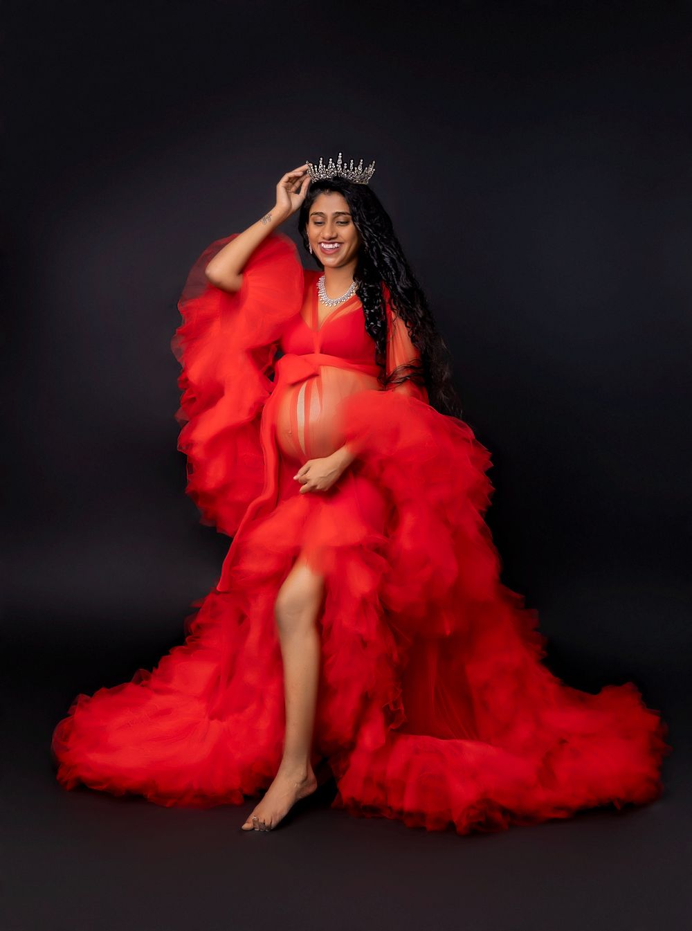 Pregnant woman in flowing red tulle gown wearing a crystal crown poses gracefully against a dark studio background.