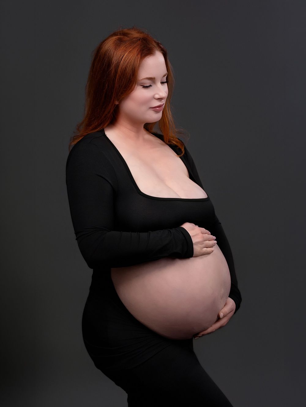 Pregnant woman in black outfit cradling bare baby bump, red hair, studio portrait on gray background.