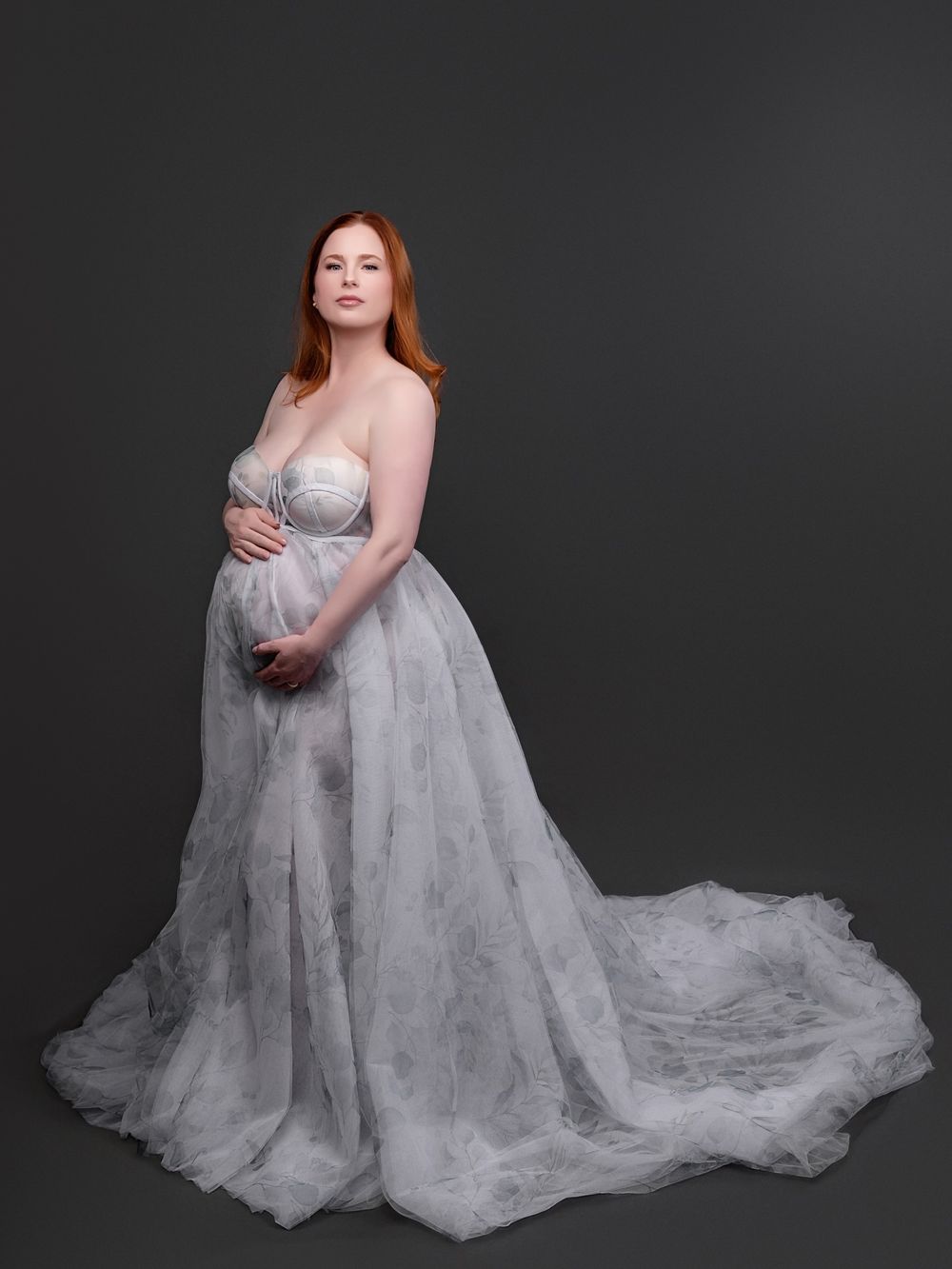 Pregnant woman in elegant strapless gray tulle gown cradling baby bump, red hair, studio portrait.