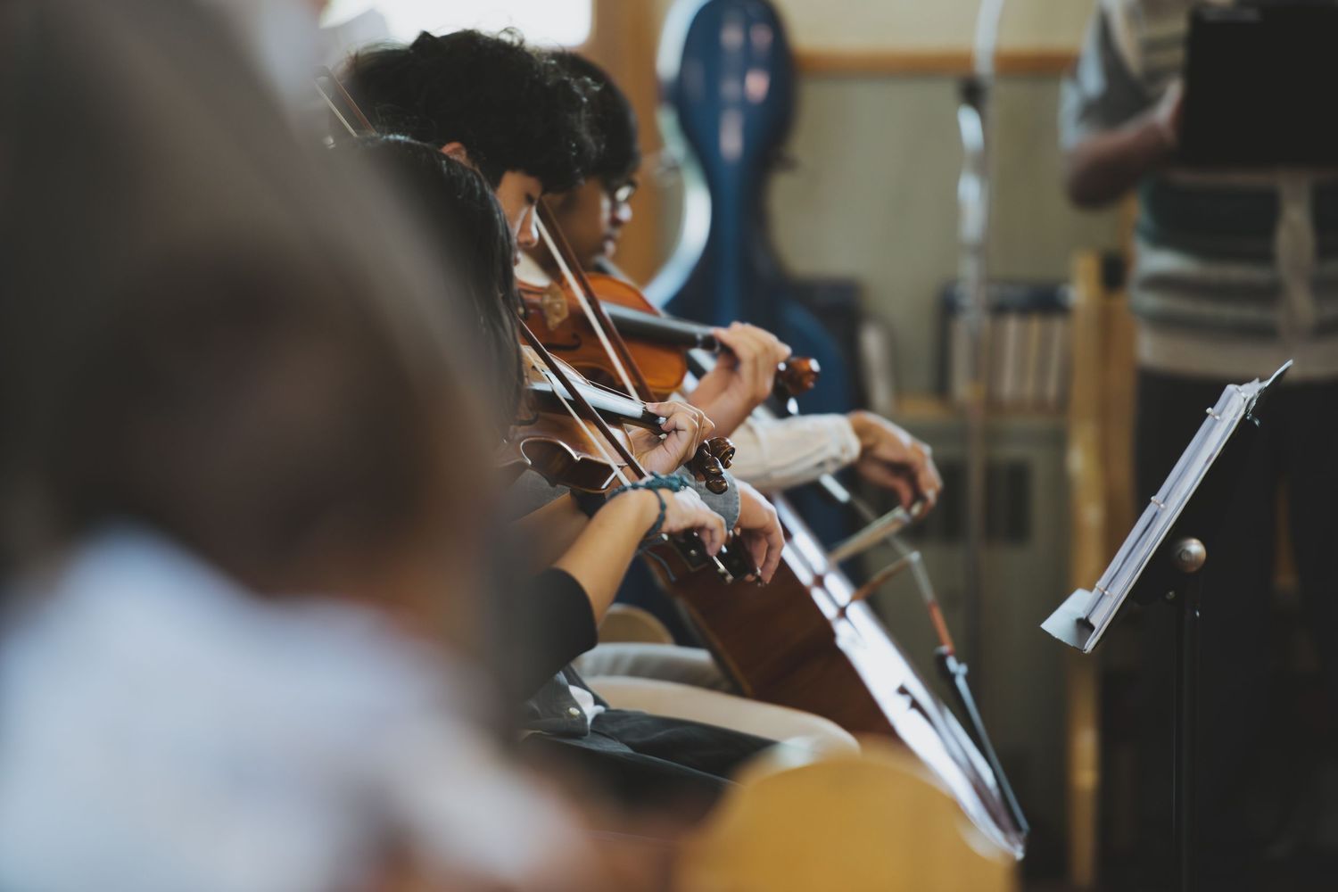 Musicians playing violins in an orchestra rehearsal, reading sheet music on stands in a warmly lit room.