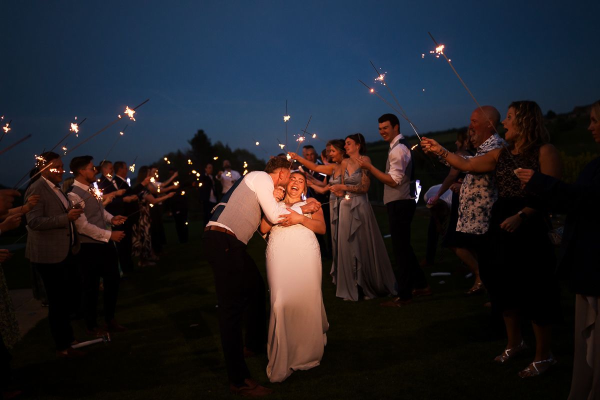 Bride and groom kissing at twilight wedding reception while guests hold sparklers in celebration outdoors.
