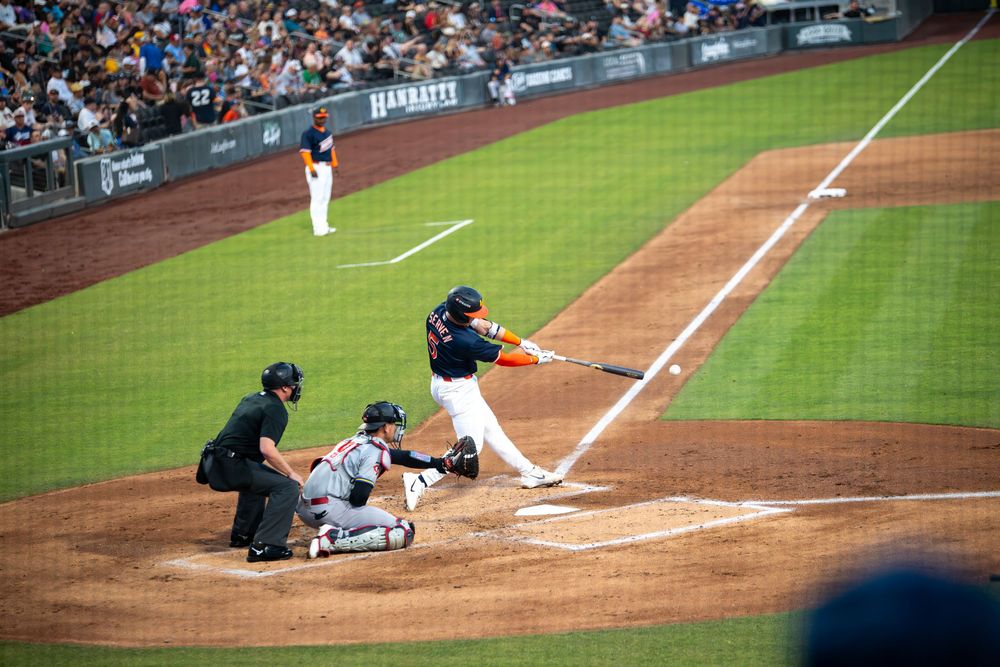 Baseball batter in orange and black uniform swinging at a pitch with catcher and umpire at home plate.
