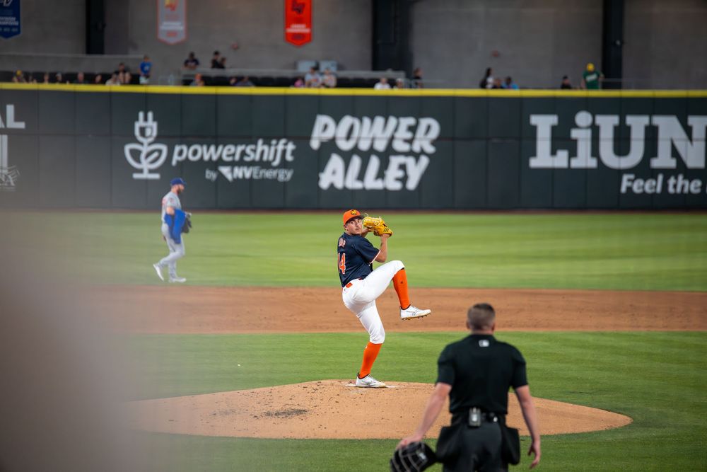 Baseball pitcher throws from mound at Power Alley stadium with umpire watching from behind.