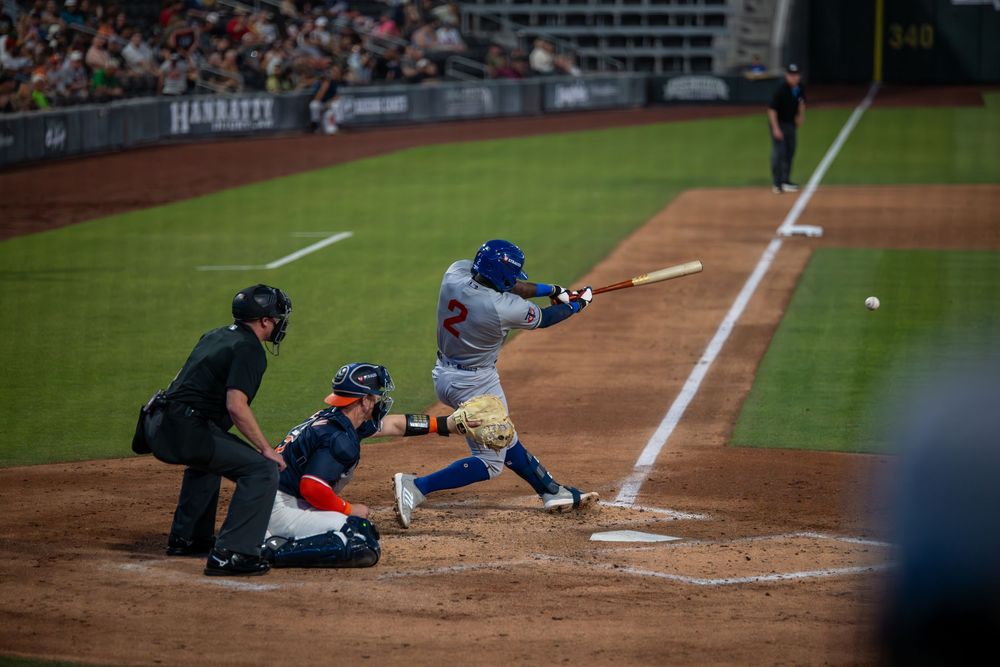 Baseball batter swings at low pitch with catcher crouching and umpire watching closely.