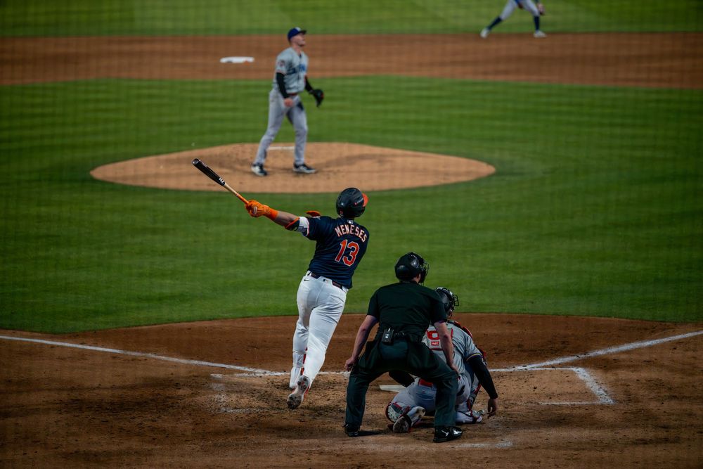 Baseball batter swings at pitch during night game with catcher and umpire in stadium.