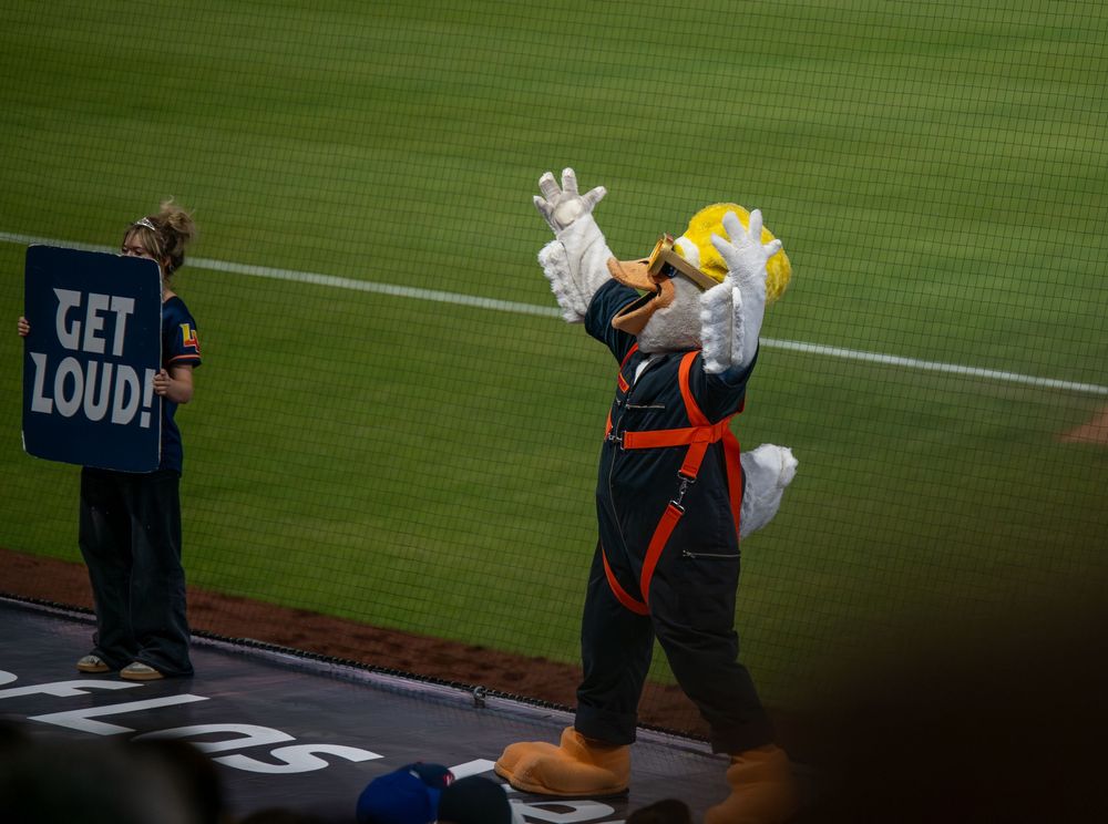 Baseball mascot in hardhat costume raises hands while fan holds GET LOUD sign on field.