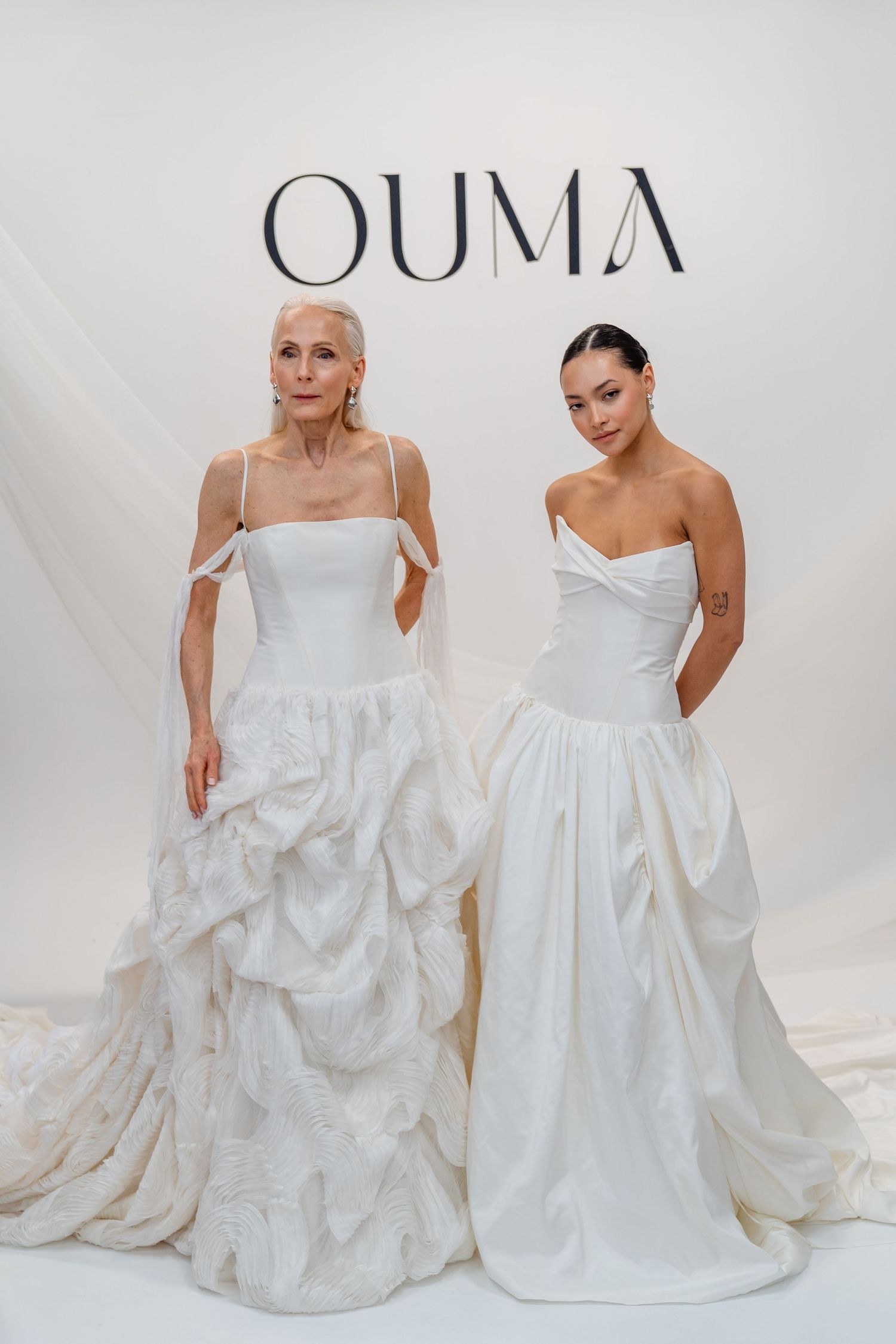 Two models wearing elegant white OUMA bridal gowns with voluminous skirts pose in front of branded backdrop.