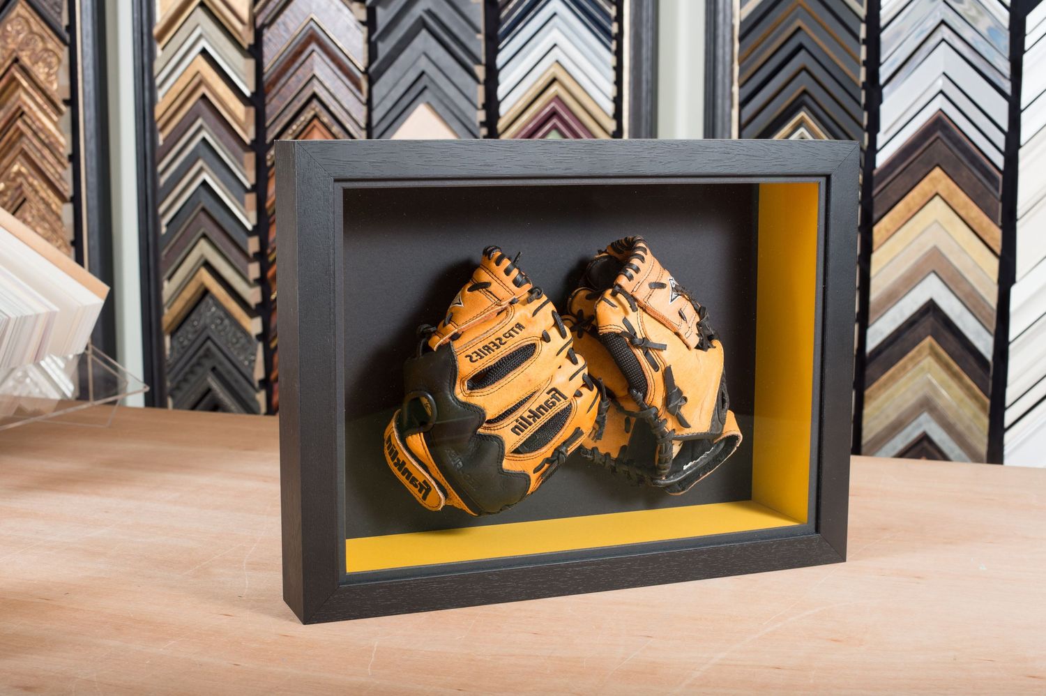 A pair of orange and black baseball gloves are displayed in a black shadow box with a yellow border on a wooden table.