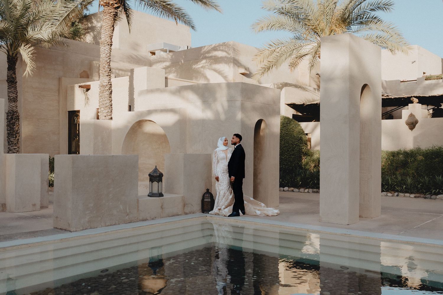 Elegant outdoor bridal portrait of a couple standing next to a serene pool inside a grand architectural resort with beautiful sunlit arches.