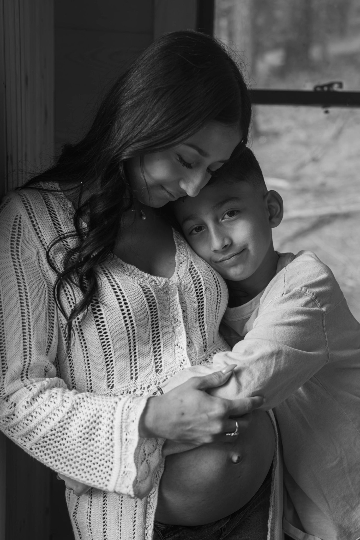 Black and white photo of a pregnant woman being hugged by her young son near a window.