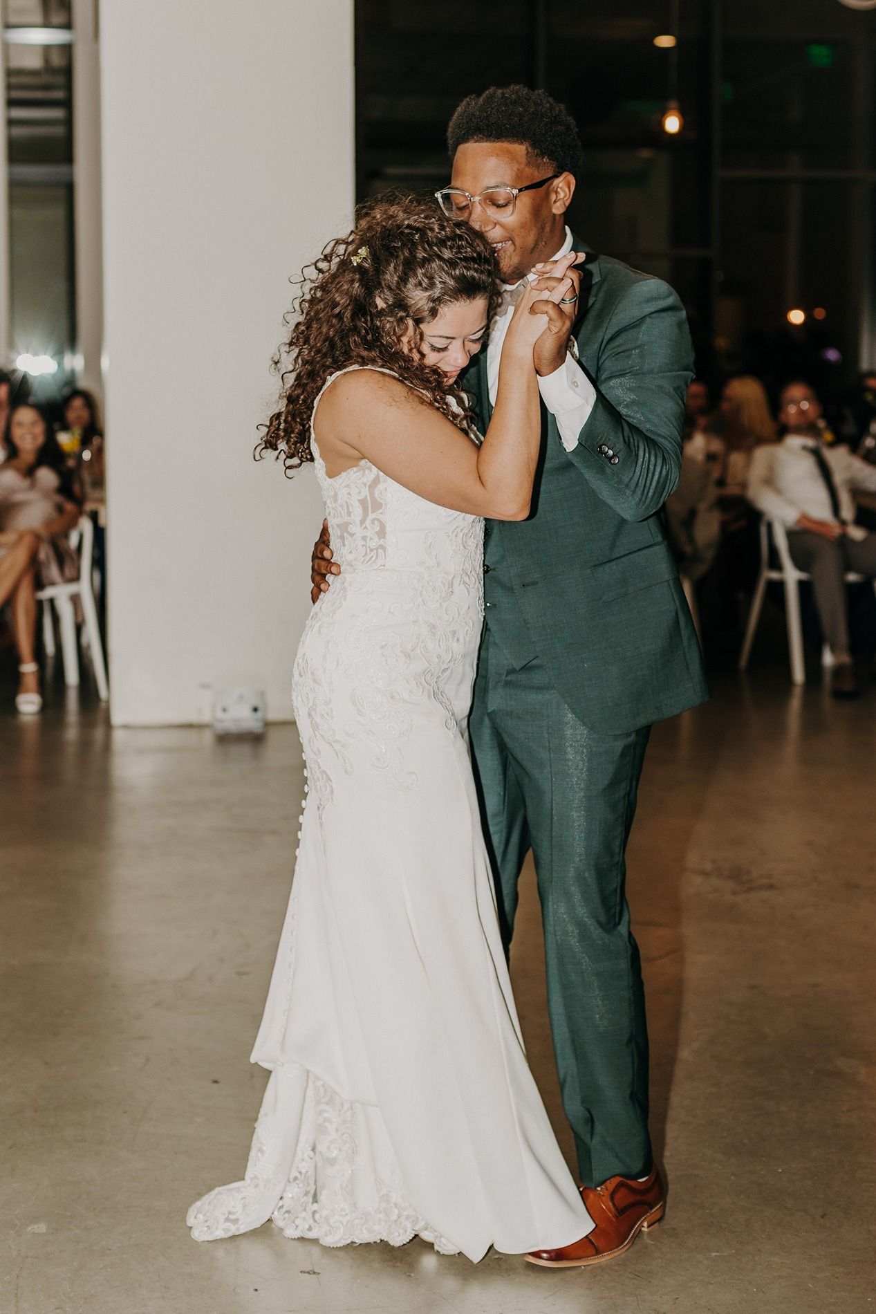 A bride and groom share their first dance, she in lace gown, he in green suit, at their wedding reception at The Lane.