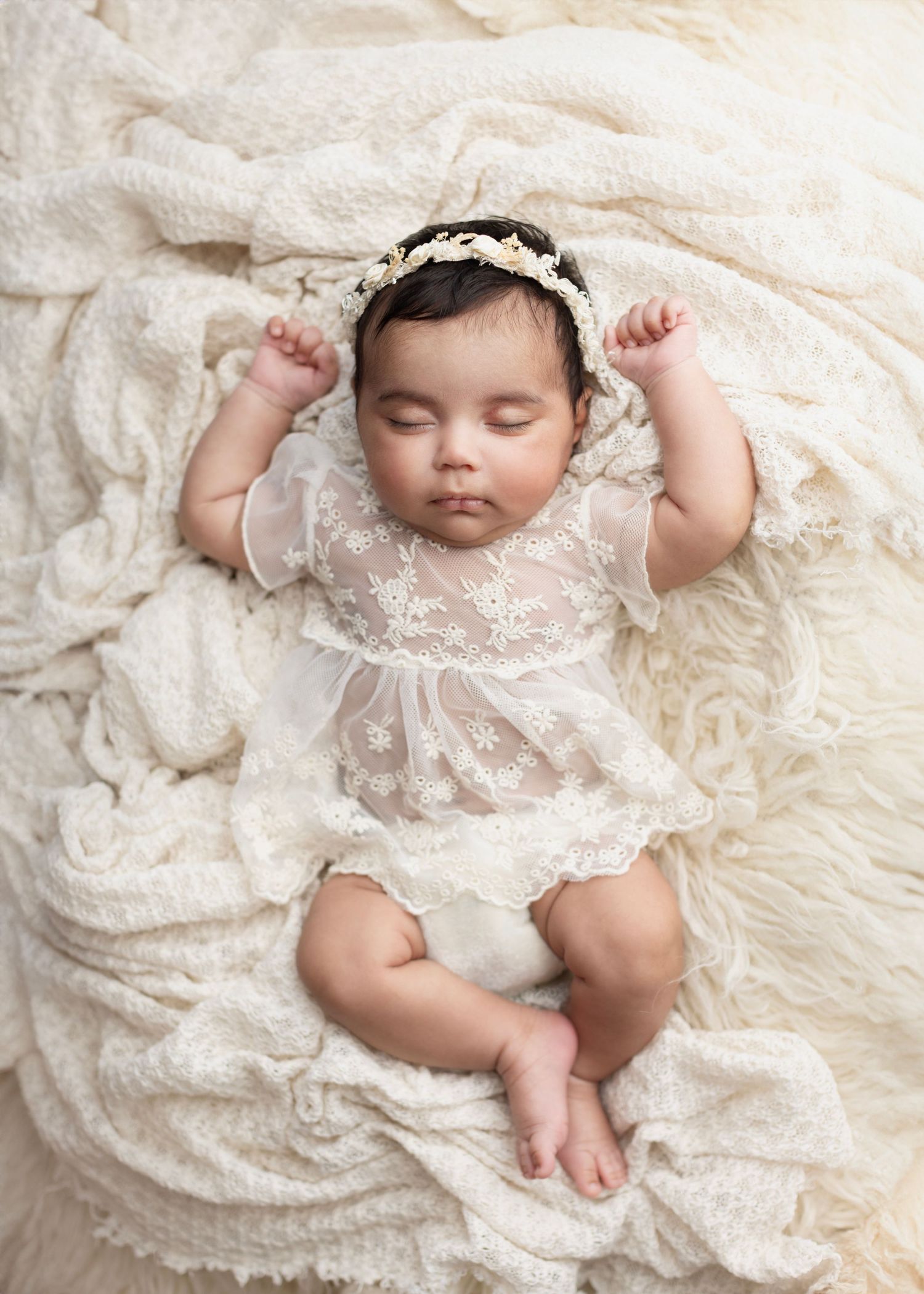 Sleeping newborn baby in white lace dress and floral headband, lying on cream knit blanket in professional portrait photo.