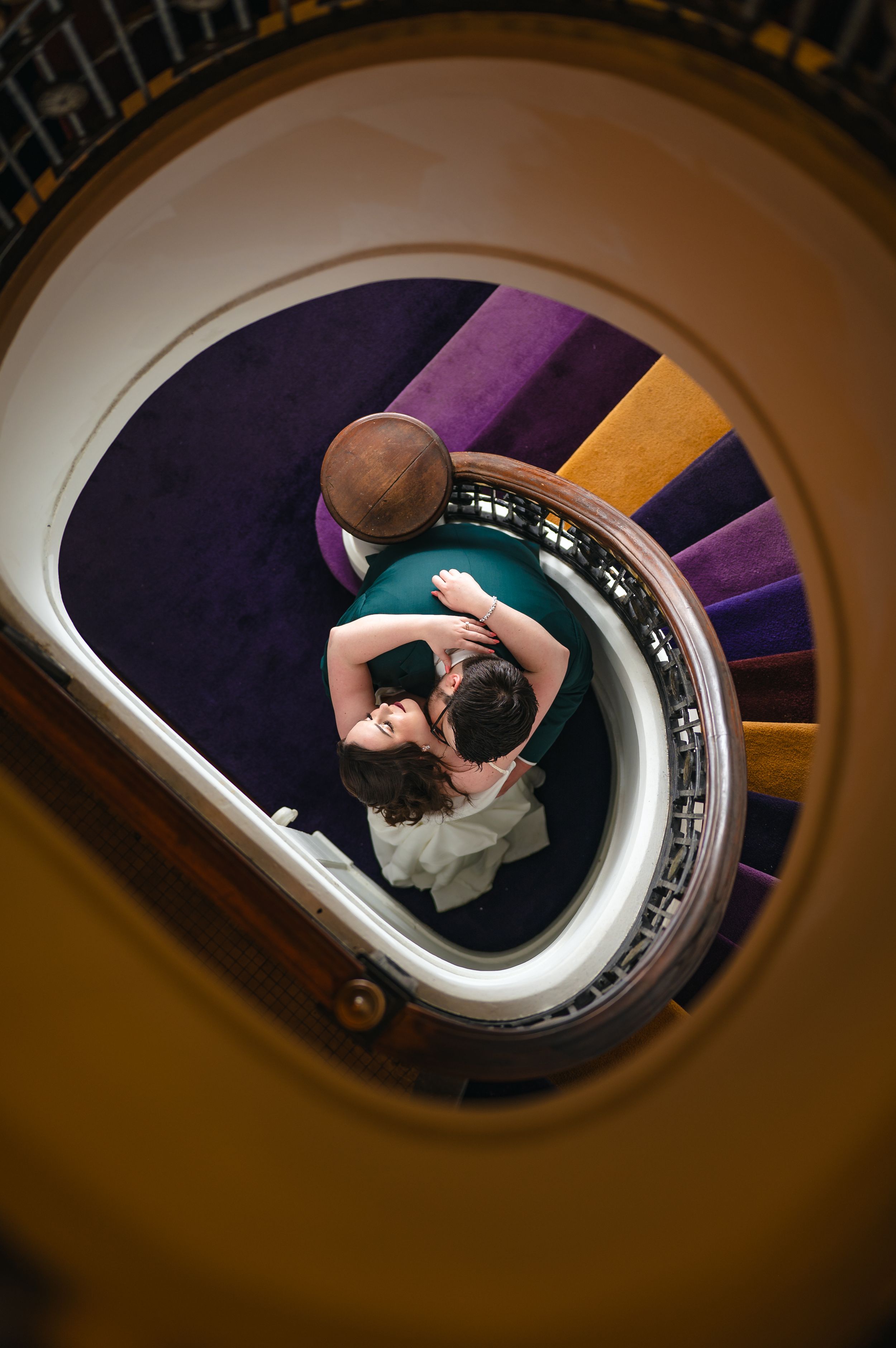Aerial view of a couple embracing on a spiral staircase with purple and gold carpet in an elegant venue.