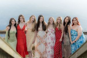 Eight young women in colorful summer dresses posing together on a wooden dock by a lake.