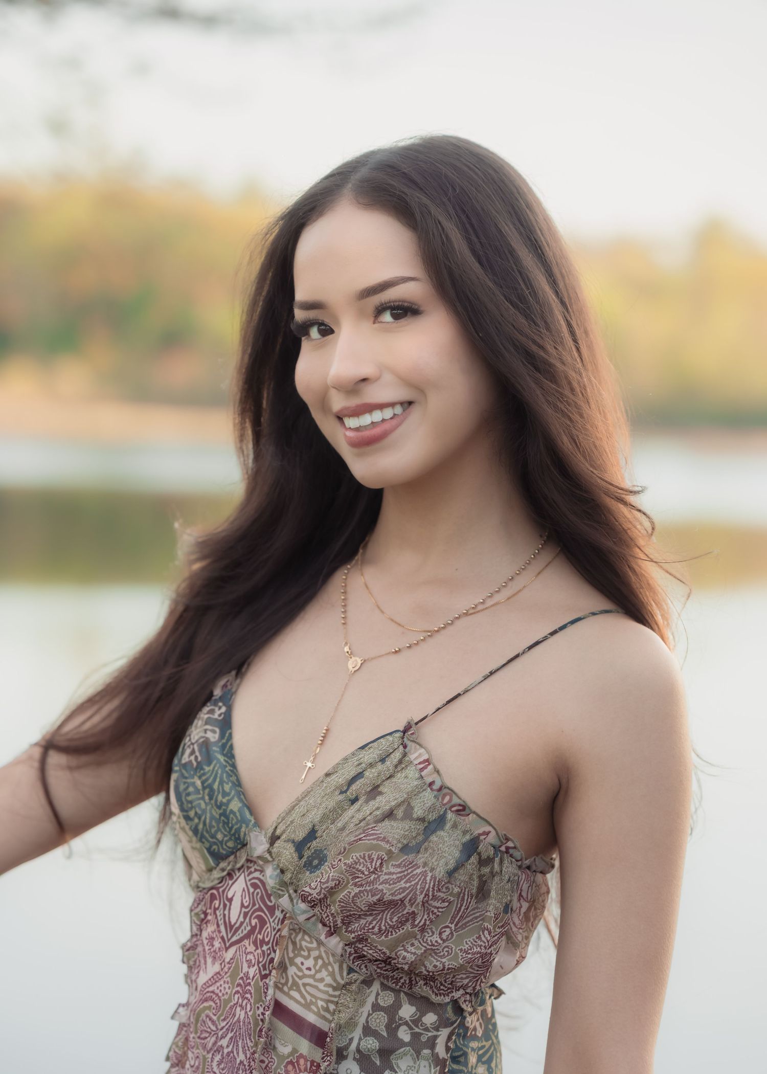 Smiling young woman with long dark hair wearing a floral sundress and gold necklace outdoors near a lake.