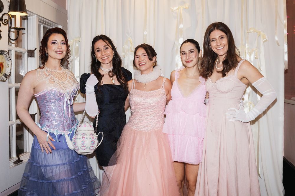 Five young women in elegant formal gowns pose together at a glamorous event, wearing pink, blue, and black dresses.