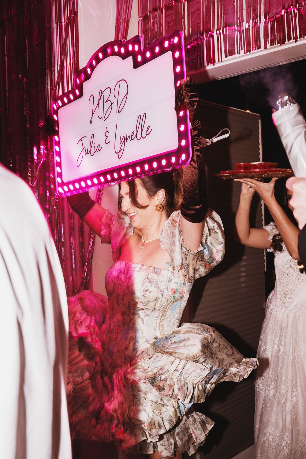 Woman in floral dress dancing under neon KBD Julia & Lynelle sign at a pink party celebration.