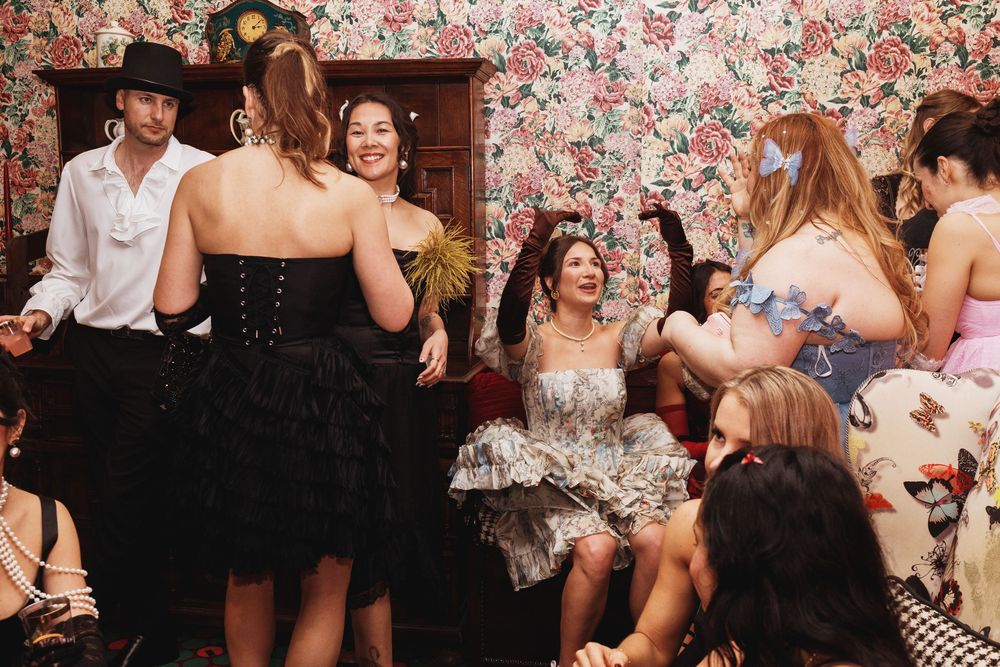 Group of women in elegant vintage dresses socializing at a floral wallpaper party venue.
