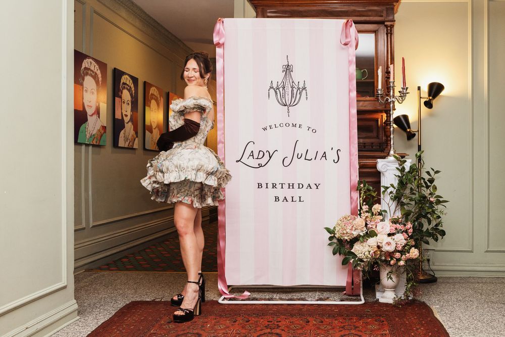 Woman in ruffled dress posing beside Lady Julia's Birthday Ball welcome sign with floral arrangements.